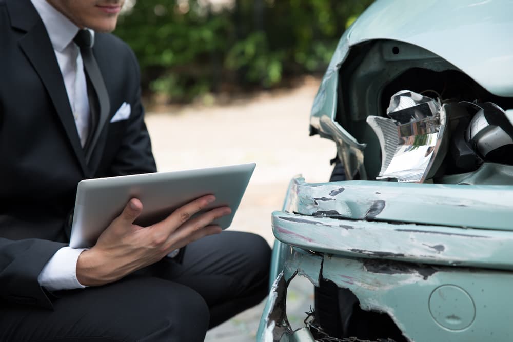 The image showing a side view of a person writing on a clipboard while an insurance agent examines a car after an accident.